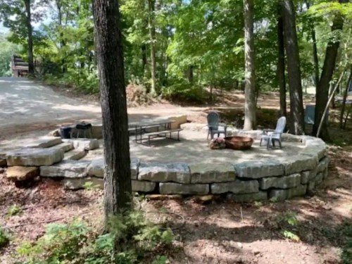 A stone fire pit area surrounded by trees, featuring chairs and a table in a wooded outdoor setting.