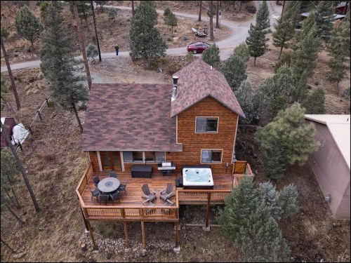 Aerial view of a wooden cabin with a deck, hot tub, and surrounded by trees in a rural area.