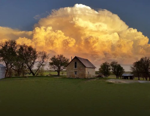 A rustic stone house sits in a green field under a dramatic sunset sky with large, fluffy clouds.