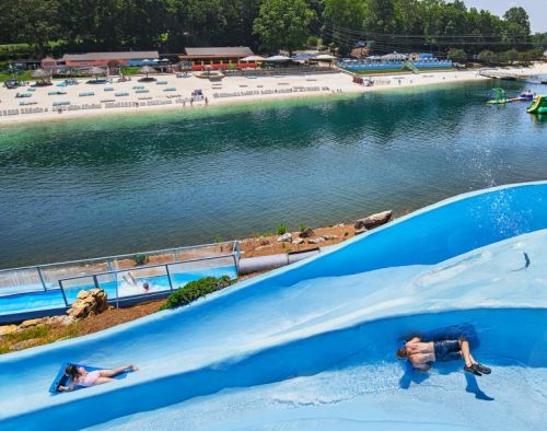 Two people slide down a blue water slide, with a beach and lake in the background on a sunny day.