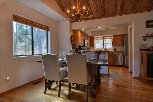 Cozy dining area with a wooden table, chairs, and a chandelier, featuring a view of trees through a large window.