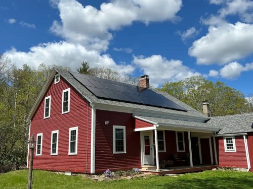 A red house with a sloped roof featuring solar panels, surrounded by greenery and a blue sky with fluffy clouds.