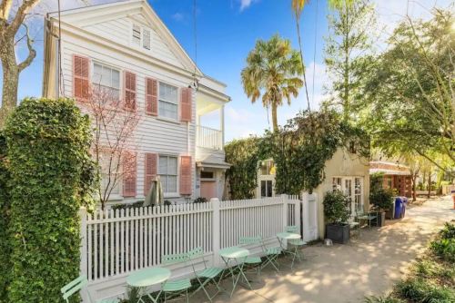 A charming white house with pink shutters, surrounded by greenery and a picket fence, featuring outdoor seating.
