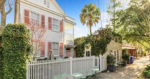 A charming white house with pink shutters, surrounded by greenery and a white picket fence, under a clear blue sky.