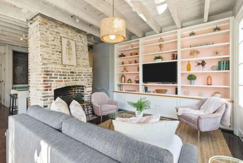 Cozy living room with a brick fireplace, pink shelves, modern furniture, and decorative plants. Bright and inviting space.