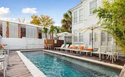 A serene pool area with lounge chairs and umbrellas, surrounded by palm trees and a white house under a clear blue sky.