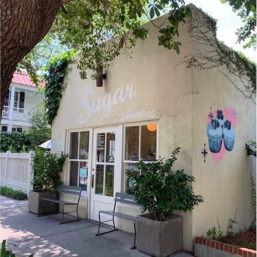 A charming bakeshop named "Sugar" with a light-colored exterior, surrounded by greenery and outdoor seating.