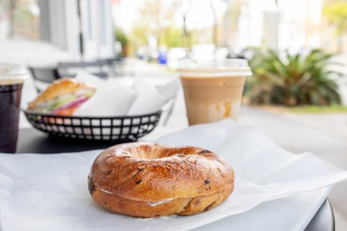 A freshly baked bagel on parchment paper, with iced coffee and a sandwich in the background.