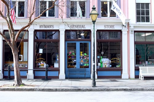 A colorful general store with large windows displaying various items, located on a street with trees and a lamppost.