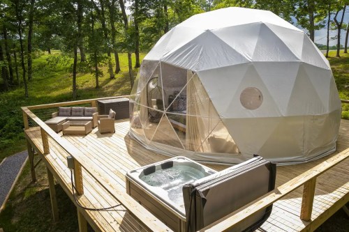 A geodesic dome on a wooden deck, featuring a hot tub and outdoor seating, surrounded by trees.