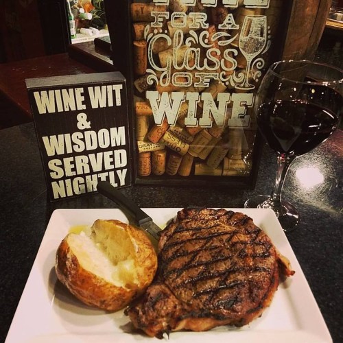 A grilled steak with a baked potato on a plate, next to a glass of red wine and a sign about wine and wisdom.