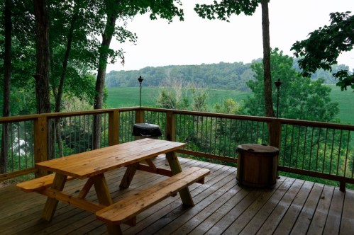 A wooden deck with a picnic table overlooks a green field and trees in a serene outdoor setting.