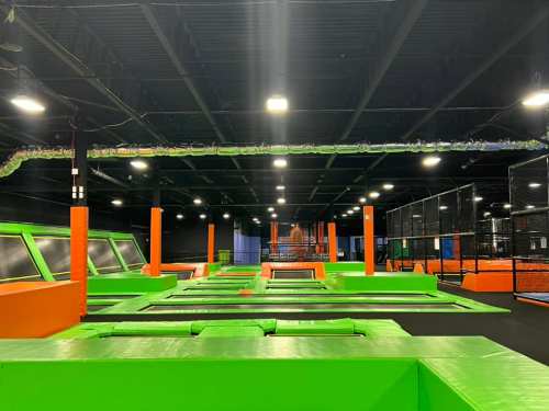 A spacious indoor trampoline park featuring green and orange trampolines and safety nets under bright lights.