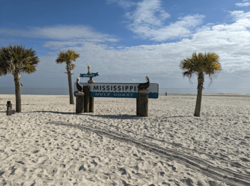 Sign for Mississippi Gulf Coast on a sandy beach, flanked by palm trees under a partly cloudy sky.