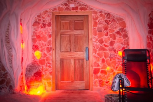 A wooden door framed by pink salt walls, with soft red lighting and a chair nearby, creating a serene atmosphere.