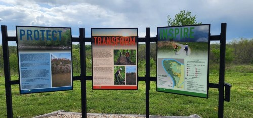 Three informational signs titled "Protect," "Transform," and "Inspire," displayed in a grassy area with a cloudy sky.