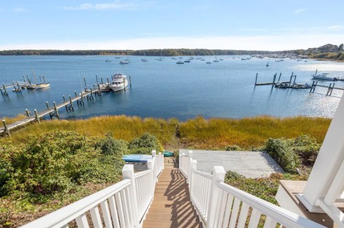 View of a serene waterfront with boats, docks, and lush greenery under a clear blue sky. Steps lead down to the water.