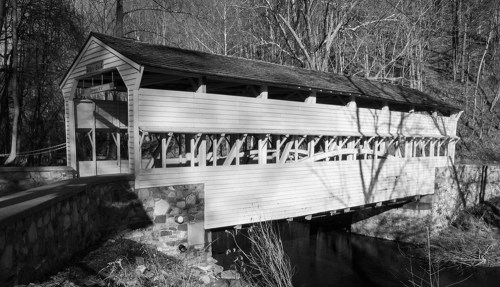 A black and white image of a covered bridge spanning a small stream, surrounded by trees.