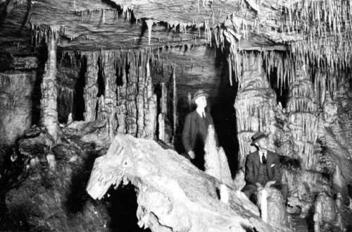 Two men in vintage attire explore a cave with stalactites and stalagmites, surrounded by rocky formations.