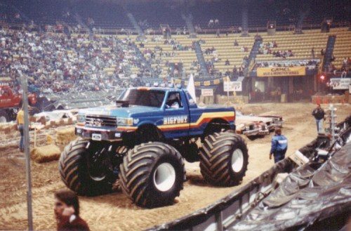 A blue monster truck with large tires drives on a dirt arena, surrounded by spectators in a stadium.
