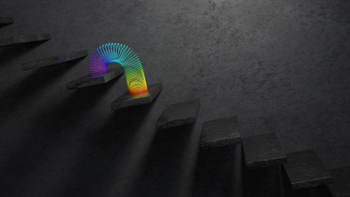 A colorful rainbow slinky rests on dark, textured steps, casting shadows in a minimalist setting.