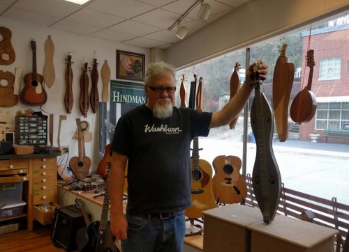 A man with glasses holds a dulcimer in a music shop surrounded by various string instruments.