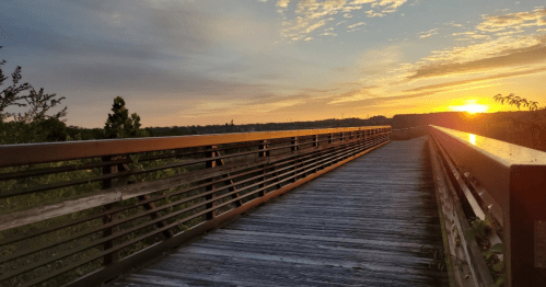 A wooden bridge stretches into the distance, framed by a vibrant sunset and a cloudy sky.