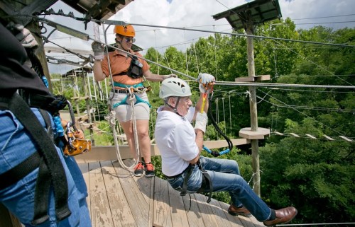 A person in a helmet zip lines while another guides them from a platform, surrounded by trees and outdoor adventure equipment.