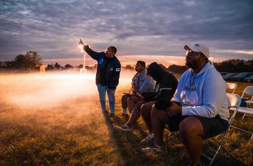 A group of four people sits on chairs in a field at dusk, with one person pointing towards the sky.