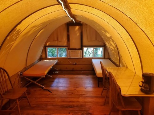 Cozy interior of a tent-like structure with two beds, a table, and an air conditioner, surrounded by natural light.