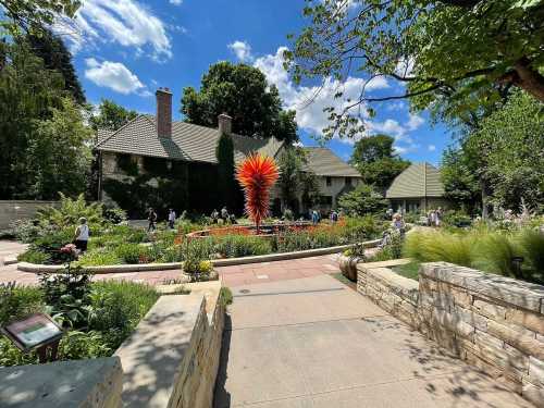 A vibrant garden with colorful flowers and a striking sculpture, surrounded by lush greenery and historic buildings.