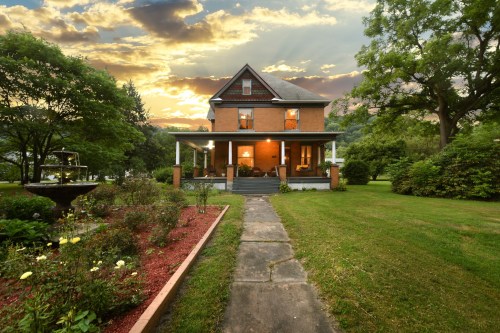 A charming two-story house with a porch, surrounded by lush greenery and a colorful garden, under a dramatic sunset sky.
