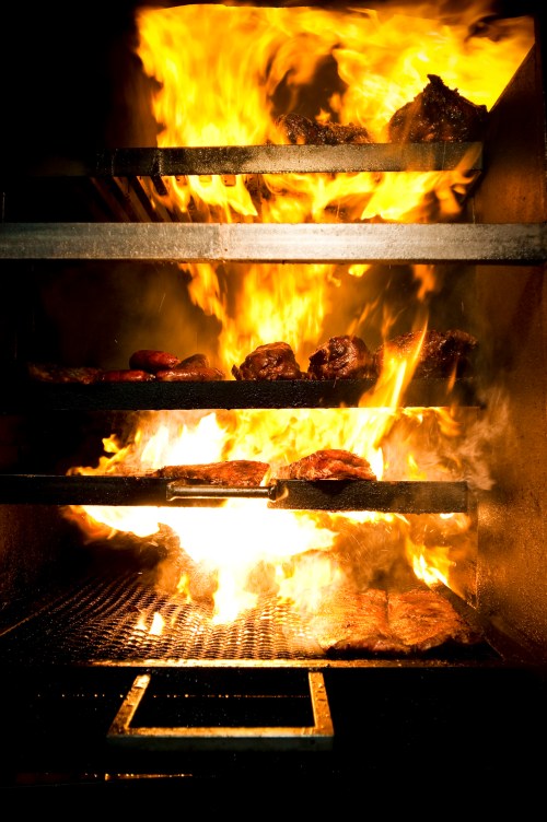 Flames engulfing a grill with various meats cooking on racks inside a smoky barbecue smoker.