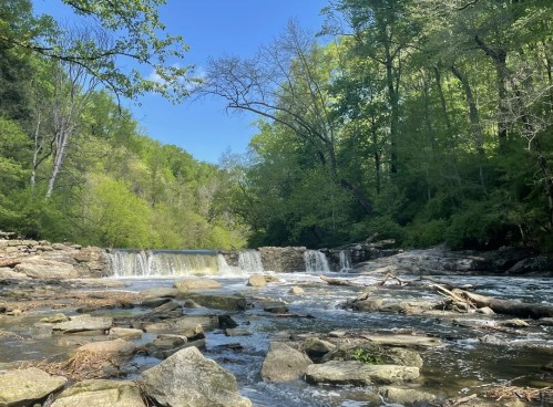 A serene river scene with a waterfall surrounded by lush green trees and rocky banks under a clear blue sky.