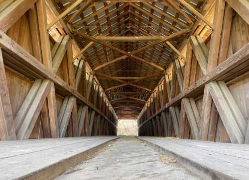 Interior view of a wooden covered bridge, showcasing its beams and open pathway leading to light at the far end.