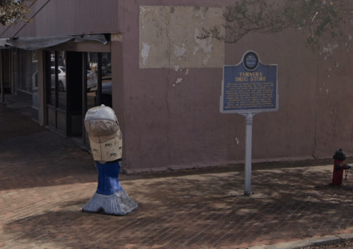 A weathered fire hydrant painted like a cartoon character stands next to a historical marker for Turner's Drug Store.