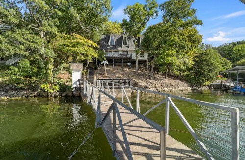 A wooden dock leads to a house surrounded by trees by a calm lake under a clear blue sky.