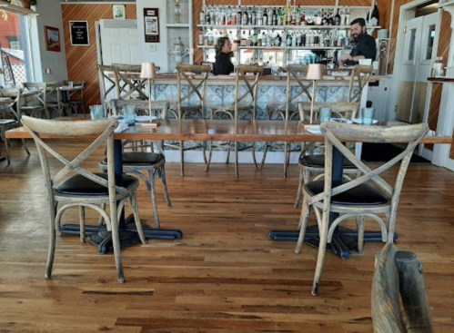 A cozy restaurant interior featuring wooden chairs, a long table, and a bar with shelves of drinks in the background.