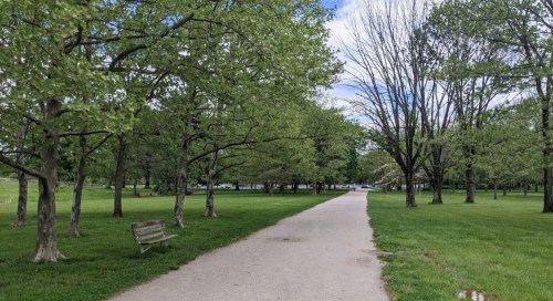 A peaceful park scene with a paved path, lush green trees, and a bench under a partly cloudy sky.