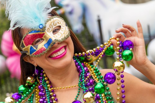A woman in a colorful mask and festive beads smiles joyfully at a celebration, with a unicorn decoration in the background.