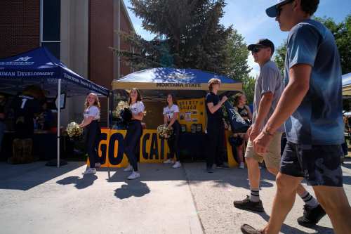 A group of students walks past a tent with cheerleaders and a "Go Cats!" sign at a Montana State event.