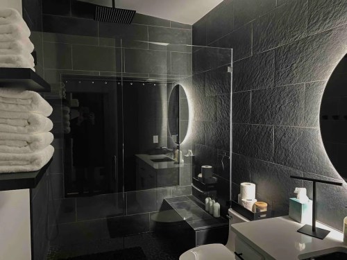 Modern bathroom featuring a glass shower, sleek black tiles, illuminated mirrors, and neatly stacked towels.