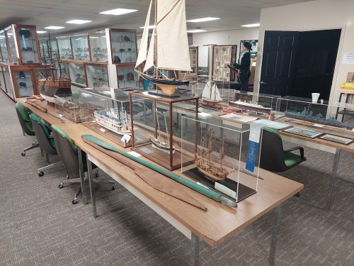 A display of model ships in glass cases on a table, with chairs and shelves of artifacts in the background.