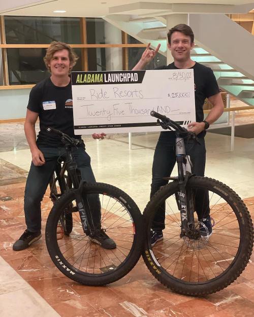 Two young men hold a large check for $25,000, standing next to mountain bikes at an event.
