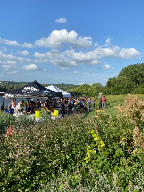 A crowd gathers near a river under a blue sky, with tents and greenery in the foreground.