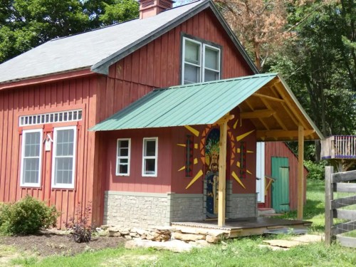 A red wooden house with a green metal roof and decorative mural on the porch, surrounded by greenery.