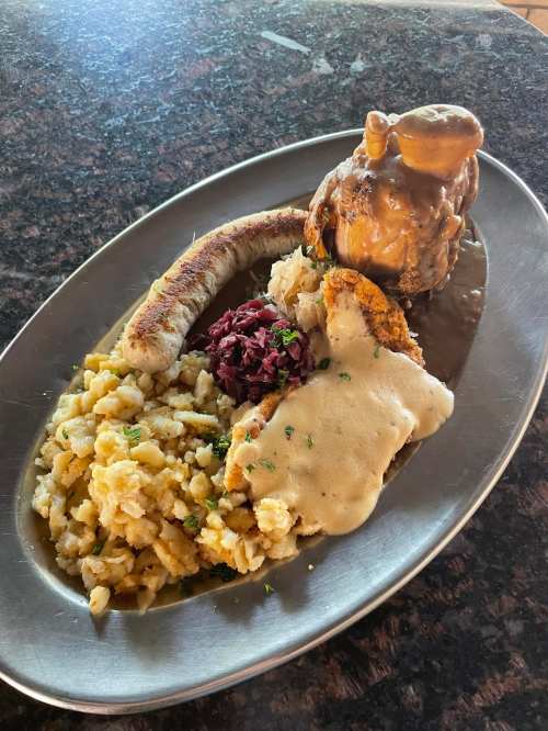 A metal platter with a hearty meal featuring sausage, fried chicken, sauerkraut, mashed potatoes, and gravy.