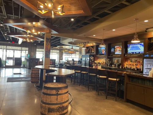 Interior of a modern bar with a wooden counter, high tables, and a well-stocked bar area, featuring TVs and warm lighting.