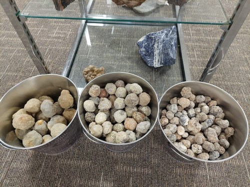 Three metal buckets filled with various round stones, displayed on a glass shelf in a museum or exhibit setting.
