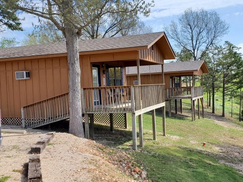 Two brown cabins on stilts surrounded by greenery, with wooden decks and a clear blue sky in the background.